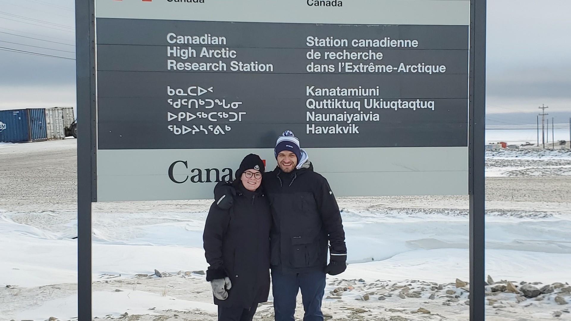 Two people in jackets standing in front of a sign in an Arctic location