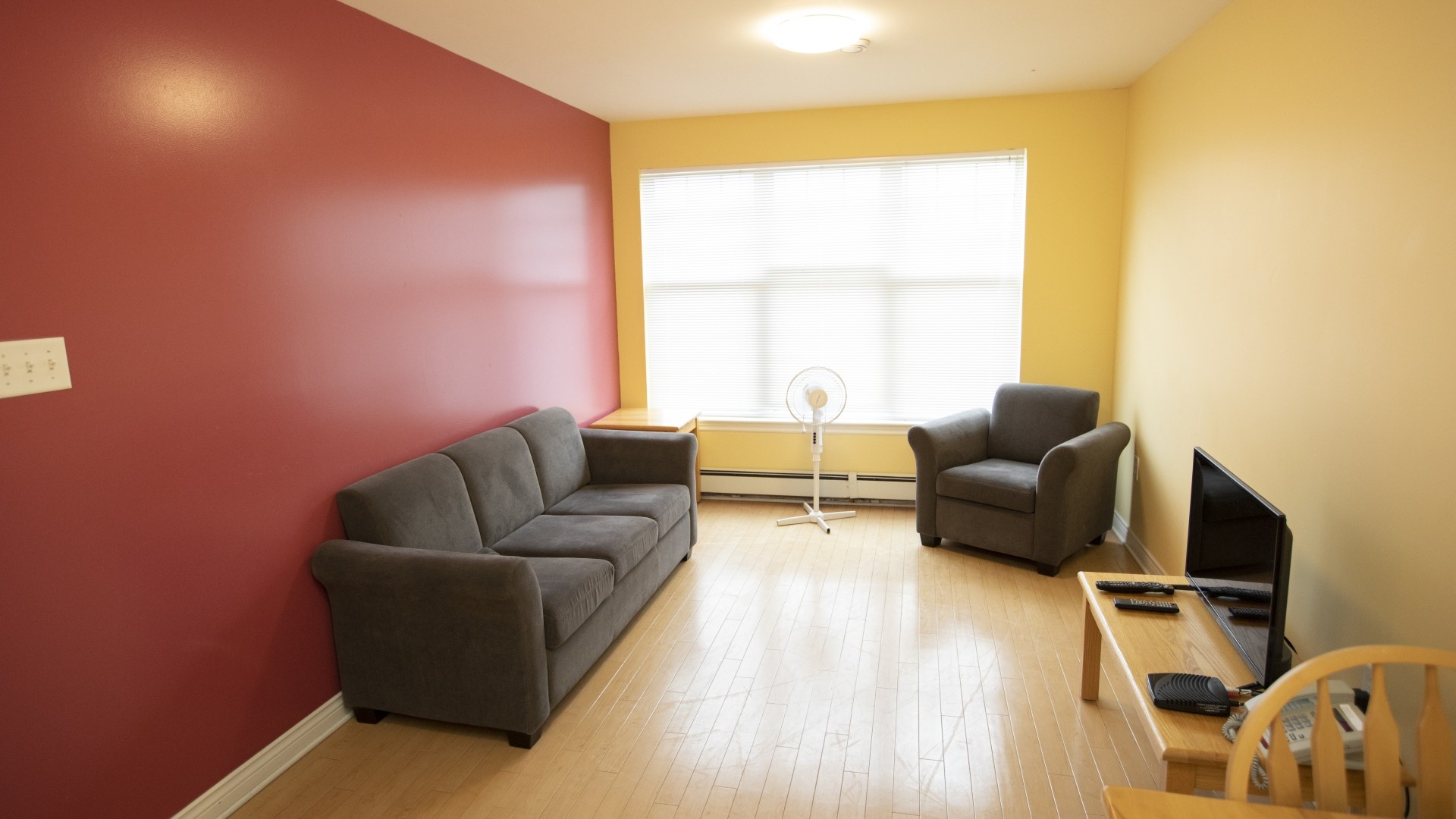 Lounge area of an apartment style room located in Powers and Somers Hall. There is a TV, along with a couch and a pedestal fan in front of the window.