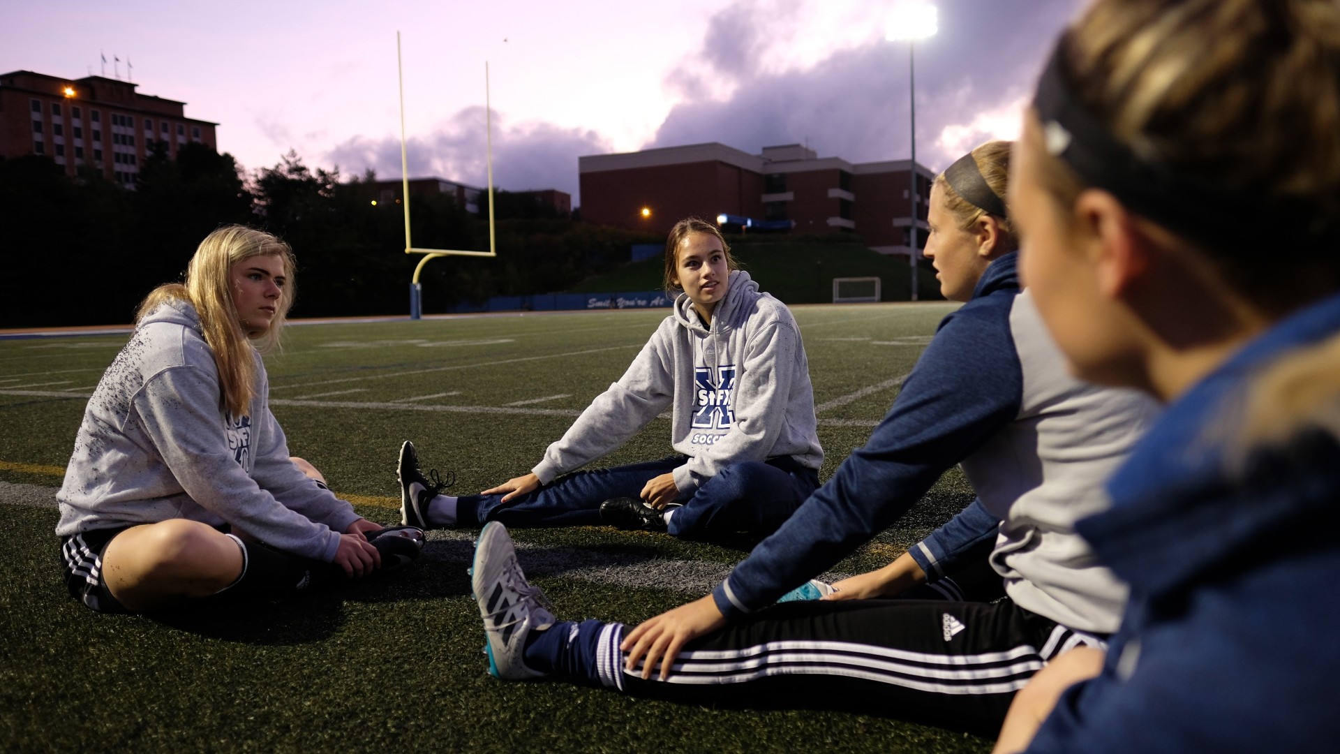 Students sitting on the football field turf to stretch during sunset.