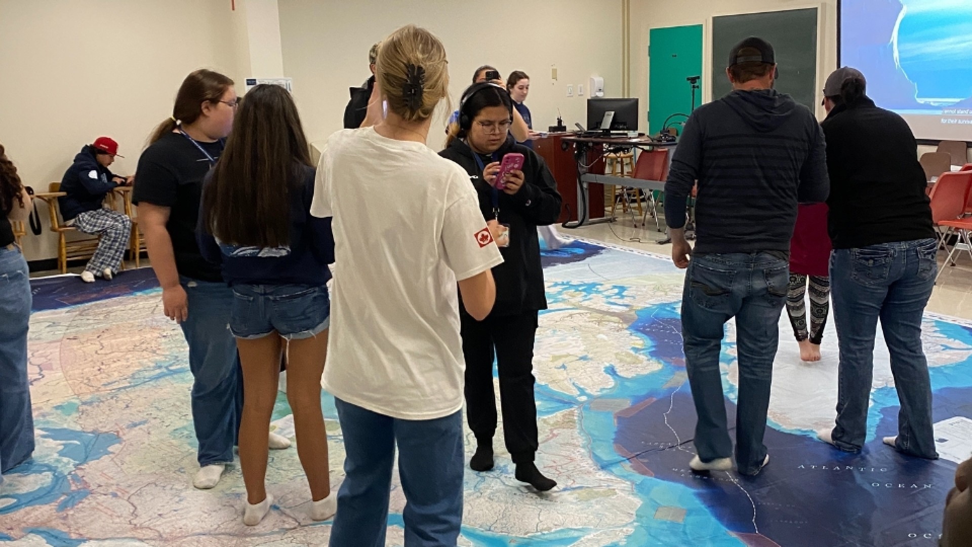 People standing on a large floor map of Canada in a classroom, engaging in an educational activity.