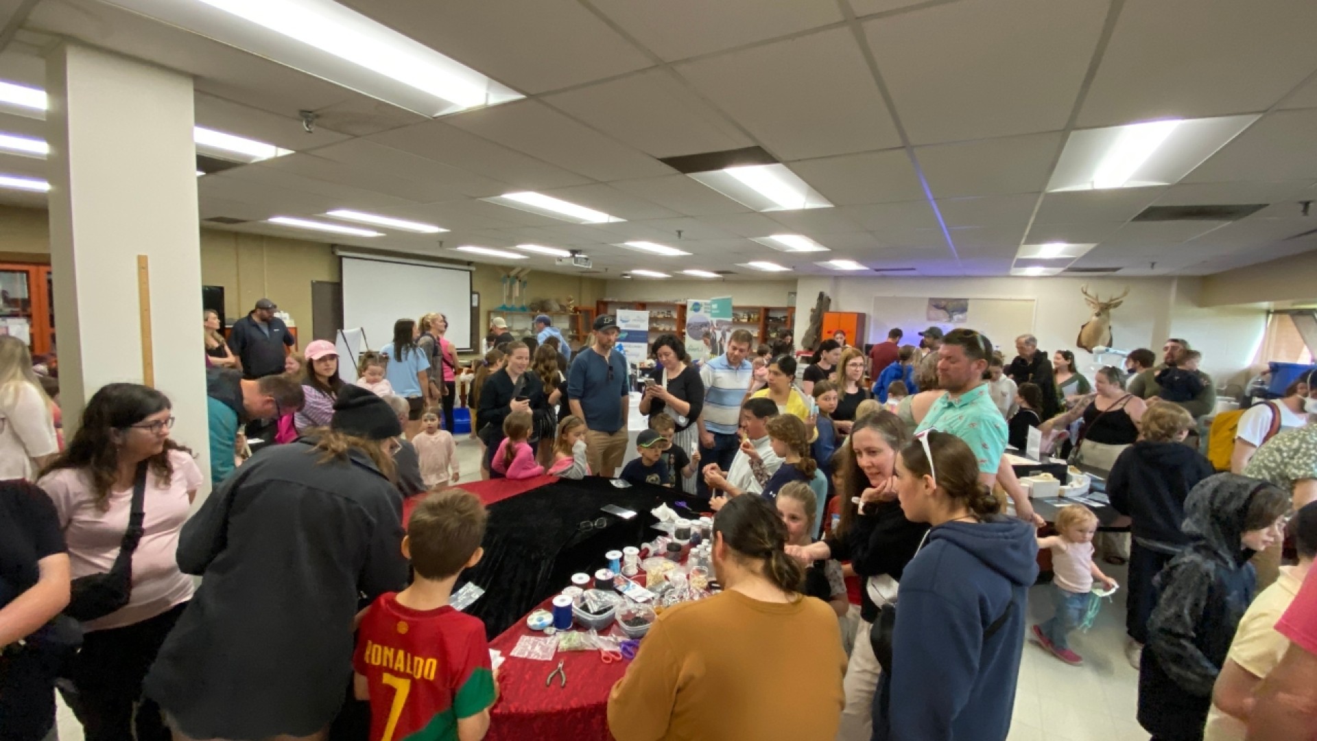 A crowded room with people gathered around tables, engaging in activities or conversations, with banners and a projector screen in the background.