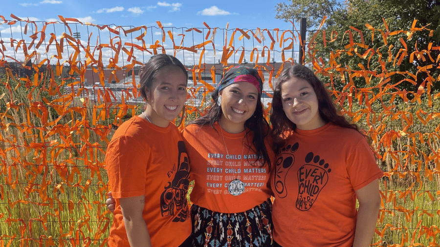 Three people wearing orange shirts in front of a field and a fence with orange ribbons