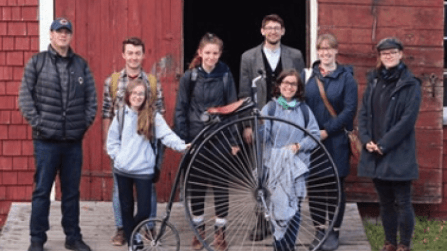 Group of people standing in front of a barn with an old-fashioned bicycle