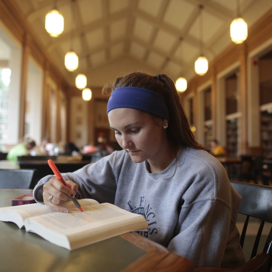 Close up of a student studying in the Angus L. MacDonald library. The student is highlighting notes in a book, while sitting at a table with other students studying in the background.
