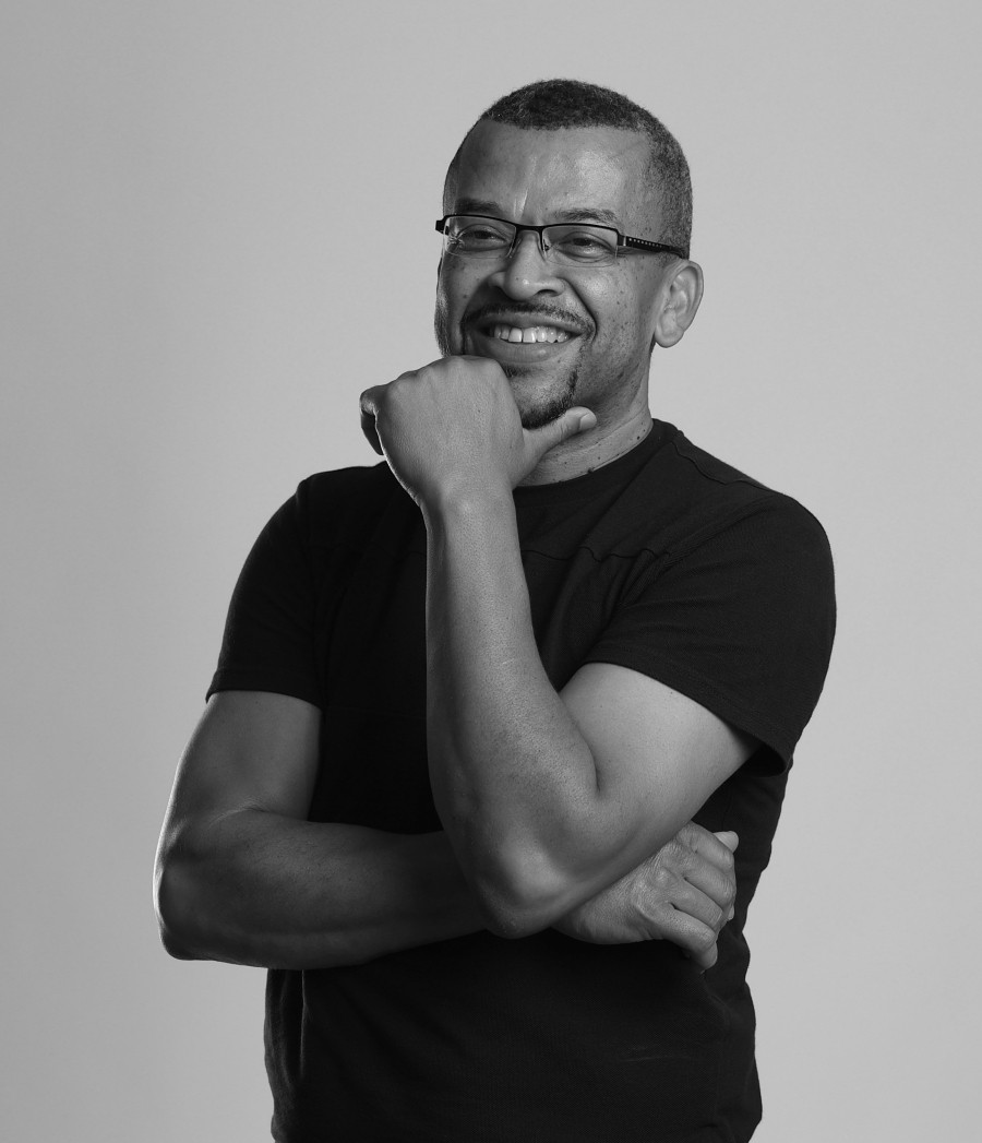 A black mid aged man stands with his arms crossed and chin resting in hand, smiling